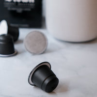 Black coffee capsule on a marble surface with a white mug and black container in the background.