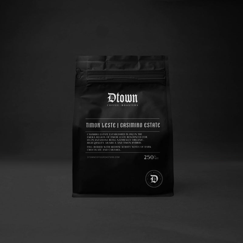 A black stand-up pouch of coffee centered against a dark gray to black studio background. The pouch has white text reading 'Blown' and 'Timor Leste i Grasimbo Estate', with a small circular 'D' logo near the bottom.